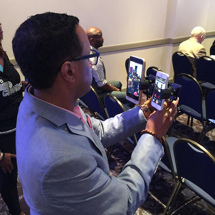A man in a light blue suit films an event with the TW Broadcaster Pro - Side-by-Side RoadVise® Tripod Mount for Live Streaming on Facebook Live, using a handheld rig in a room with rows of blue chairs and several seated people.