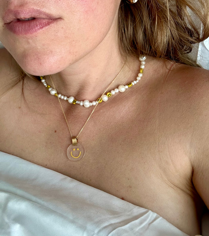 A close-up image shows a person wearing two necklaces. One necklace features pearls and smiley face beads, while the other is WALD Berlin's Smilie Dude Medaillon Gold Necklace, an 18k gold-plated piece with a transparent pendant made in Germany. The person is wrapped in a white sheet.