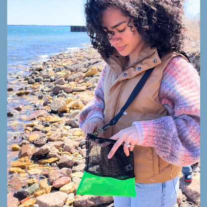 Woman on a rocky beach holding a black mesh bag with green trim. Hands-free mesh rock collecting bag with included carrying strap for Lake Superior agates, hiking, beachcombing, and foraging. AdventureUs Mesh Collecting Bag