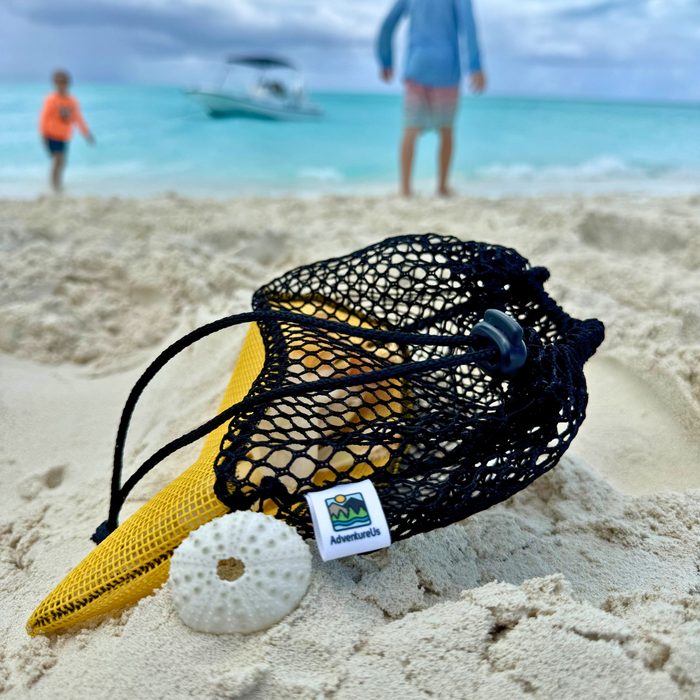 Yellow and Black mesh bag on a sandy beach with people and a boat in the background. AdventureUs Mesh Collecting Bag. Mesh collecting bag used for rocks, shells, mushroom and berry foraging, and outdoor nature treasures. AdventureUs Mesh Collecting Bag