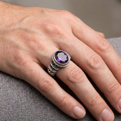 Close-up of a man's hand wearing a 925 sterling silver spine ring featuring a large, faceted oval purple amethyst gemstone, resting casually on a textured grey fabric.