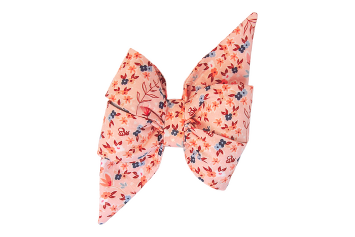 Feminine fabric dog belle bow in a blush pink shade, decorated with a dense pattern of small flowers interspersed with tiny bee illustrations and leaf motifs, tied into two full loops with a central knot and two elongated, pointed tails.