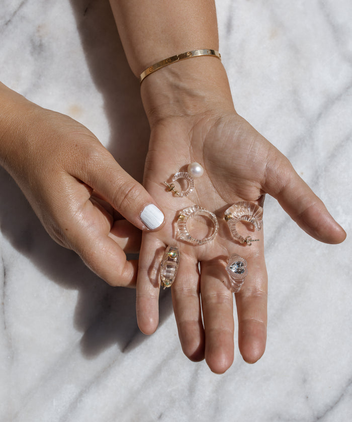 A hand with white nail polish showcases the WALD Berlin Lover Transparent Ring alongside clear and gold earrings, including hoops, a pearl stud, and a Swarovski-accented piece, all arranged on a marble surface.