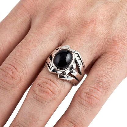 Close-up of a man's hand wearing a chunky sterling silver rocker ring featuring a large oval black onyx stone and carved band details, set against a plain white background.
