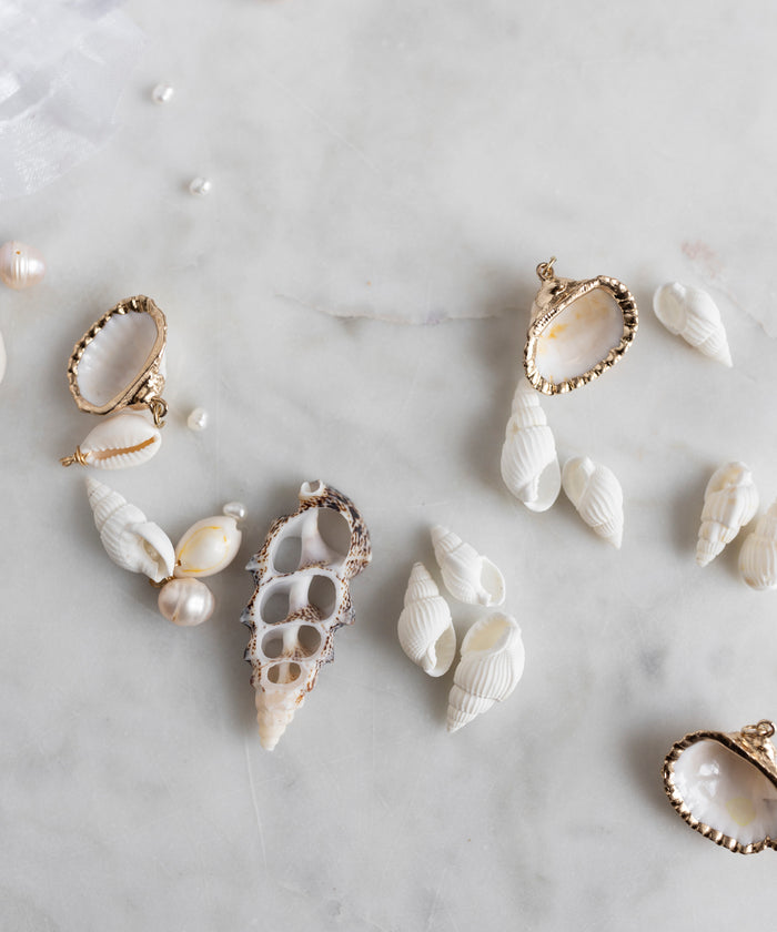 The WALD Berlin Candyman Shell Kit features handpicked, assorted white seashells and elegant gold-edged shell pendants artfully displayed on marble, each shell standing out as a unique jewel piece.