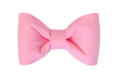 A dog bow tie made from soft solid pink fabric, featuring a classic double-loop design with wide, rounded ends and a central cinch. The material has a smooth texture and subtle luster, suggesting comfort and a stylish look when attached to a collar.