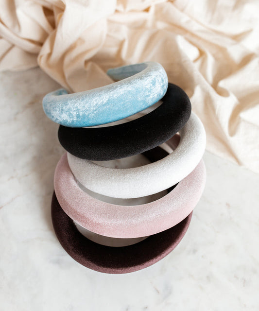 Five WALD Berlin Classic Rose Velvet Headbands in pastel shades of blue, black, white, pink, and burgundy are stacked on a marble surface with a beige cloth in the background—a touch of elegance made in Germany.
