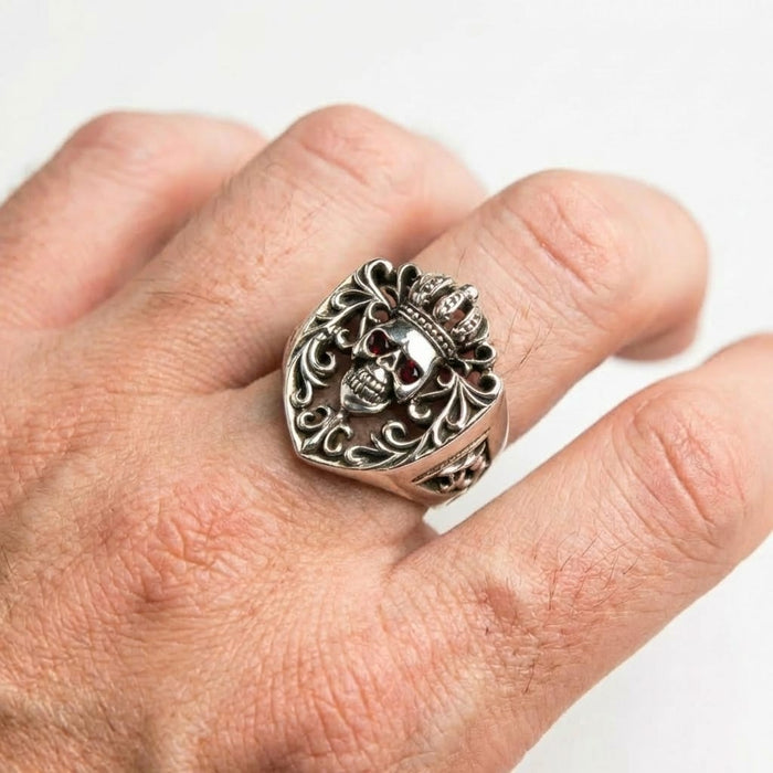 Close-up of a man's hand wearing a gothic sterling silver ring featuring a crowned skull with red stone eyes and intricate filigree details, set against a plain white background.