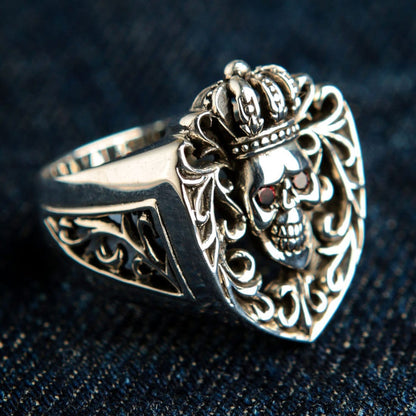 Silver shield-shaped ring face resting on denim, showcasing the crowned skull, red garnet CZ eyes, and gothic openwork.