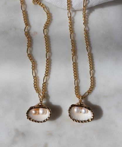 Two WALD Berlin Drop It Like It's Hot Shell Necklaces, gold-plated and handmade with shell pendants holding two small pearls each, are displayed on a light marble surface—a modern spin on the classic pearl necklace.
