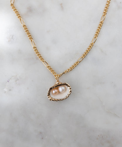 The Drop It Like It’s Hot Shell Necklace by WALD Berlin features a gold-plated pendant with two pearls in a textured oval frame, displayed on white marble—a stunning piece of handmade jewelry.