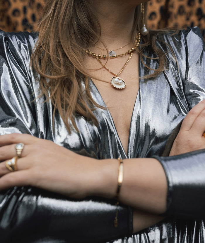 Wearing a metallic silver long-sleeve dress and layered WALD Berlin's handmade gold jewelry with pearl accents, including the Drop It Like It’s Hot Shell Necklace, arms crossed, against a leopard print background.
