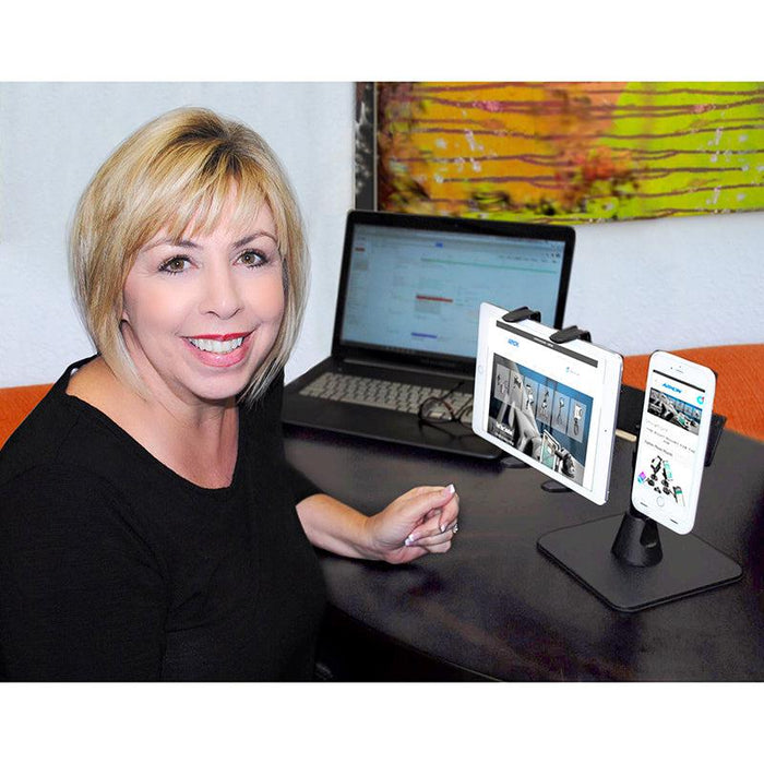A woman uses the Dual Slim-Grip® Tablet Holder and RoadVise® Phone Holder with Desk Stand Mount for Side-by-Side Streaming at her desk, holding her tablet and phone side by side, with different content on each screen and art in the background.