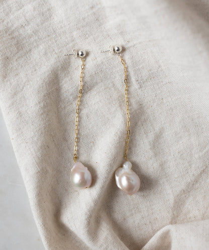 The WALD Berlin Happy Holidays Pearl Drop Earrings feature gold chains and irregular-shaped pearl pendants, shown on a beige fabric background.