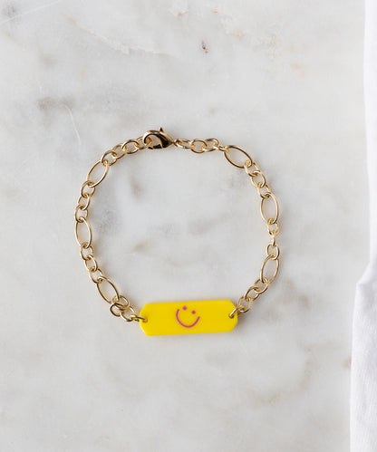 The WALD Berlin SAMPLE SALE Happy ID Yellow bracelet exclusive features a yellow tag with a pink smiley face, made in Germany, shown on a white marble surface.