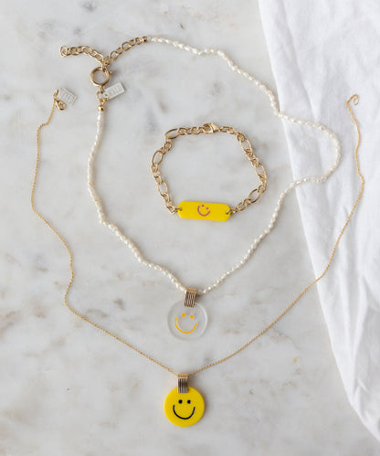 On a marble surface: WALD Berlin SAMPLE SALE Happy ID Yellow bracelet exclusive with pearls and 18k gold plating, plus two necklaces—one pearl and gold with a clear smiley pendant, one gold with a yellow smiley pendant.