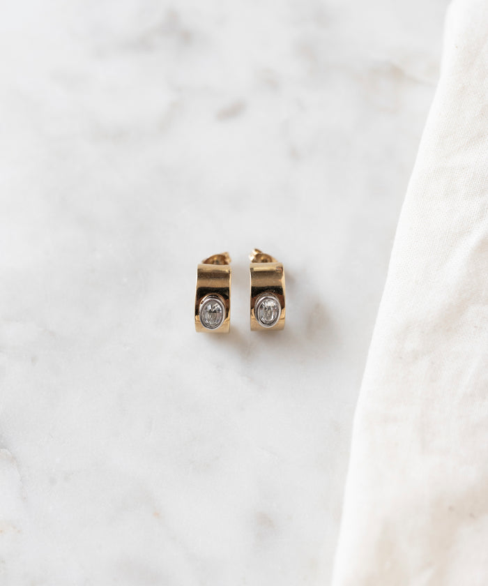 The Isla Bonita just a friend earrings by WALD Berlin, delicate stud earrings crafted from recycled gold and adorned with clear gemstones, are elegantly displayed on a marble surface beside a white cloth.
