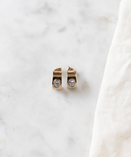 The Isla Bonita just a friend earrings by WALD Berlin, delicate stud earrings crafted from recycled gold and adorned with clear gemstones, are elegantly displayed on a marble surface beside a white cloth.