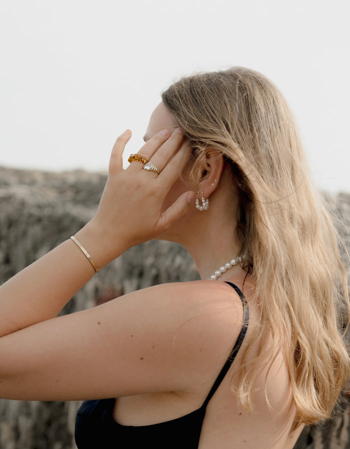 A woman with long blonde hair and a black top stands outdoors, facing away, touching her face. She wears WALD Berlin's Isla Bonita Pearl Hoops, a pearl necklace, and gold jewelry.