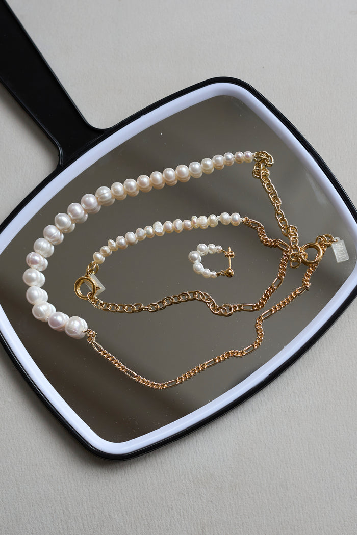 A handheld mirror displays WALD Berlin’s Isla Bonita Pearl Hoops, a pearl necklace, a pearl bracelet with a gold cross charm, and two gold plated sterling silver chain necklaces.
