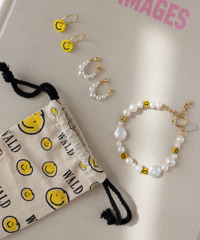 WALD Berlin's Isla Bonita Pearl Hoops, sweetwater pearl earrings, a pearl and smiley face bracelet, and a drawstring pouch with smiley faces are displayed on a beige surface.