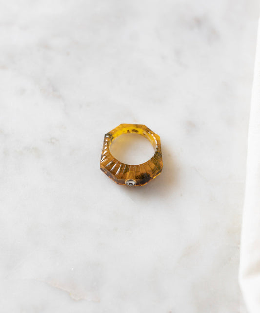 An Isla Bonita Shining Star ring by WALD Berlin, featuring a hexagonal, amber-colored gemstone, lies on a light marble surface.
