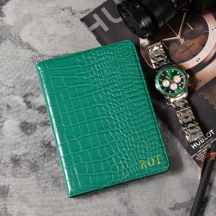 Green crocodile-patterned wallet with 'ROT' initials on a gray surface with a Hublot watch and camera.