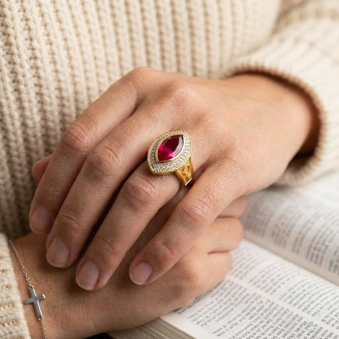 Hands resting on an open book modeling the gold-plated marquise ruby ring, highlighting the sparkling halo setting.