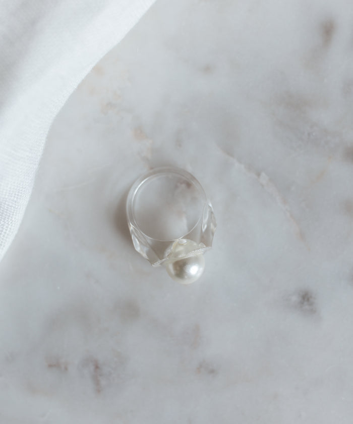 The WALD Berlin Happy No No No Ring SAMPLE, with its translucent band and Swarovski pearl, adorns a marble surface partially covered by white fabric.