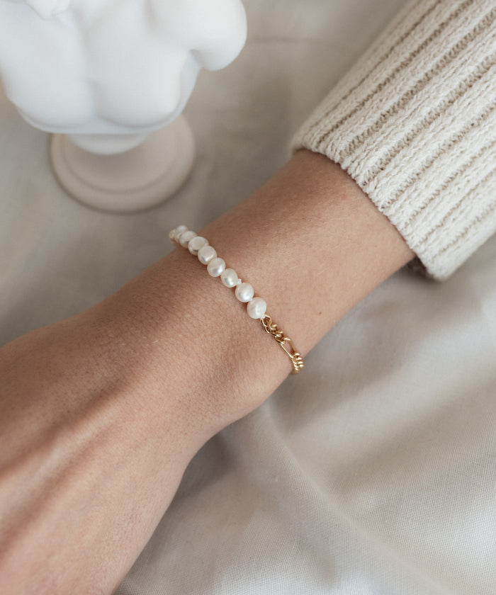 A person wearing the WALD Berlin Pas De Deux bracelet/anklet alongside a gold chain bracelet, with part of a cream-colored long sleeve visible.