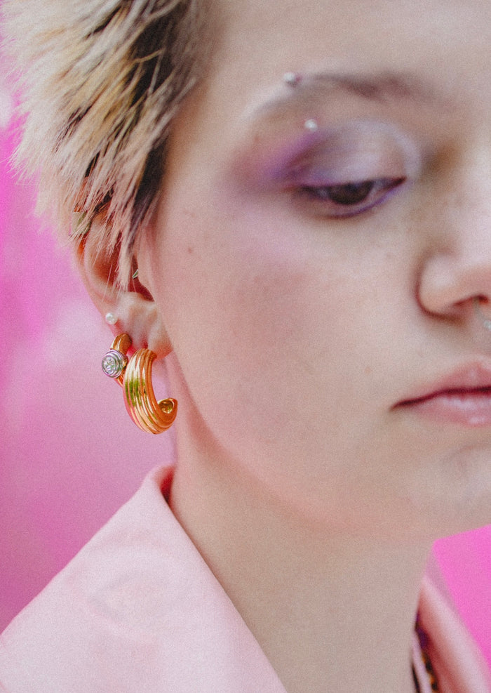 A person with short, light hair wears WALD Berlin’s SAMPLE SALE Pop Bi Color Crystal Earrings, a pink shirt, and purple eyeshadow, set against a pink background.