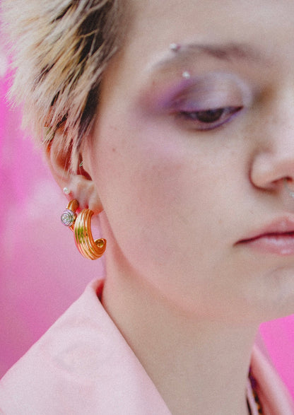 A person with short, light hair wears WALD Berlin’s SAMPLE SALE Pop Bi Color Crystal Earrings, a pink shirt, and purple eyeshadow, set against a pink background.