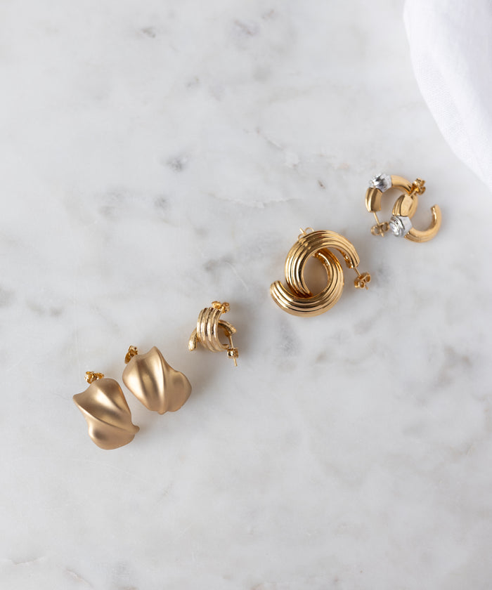 Four pairs of WALD Berlin Pop Wave Earrings Big in gold-plated brass are arranged on white marble, showcasing abstract styles, creoles, knotted designs, and gem-set hoops—all with comfortable 925 silver pins.
