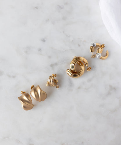 Four pairs of WALD Berlin Pop Wave Earrings Big in gold-plated brass are arranged on white marble, showcasing abstract styles, creoles, knotted designs, and gem-set hoops—all with comfortable 925 silver pins.