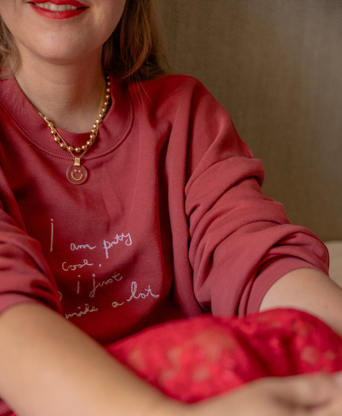A person wearing a limited edition PRETTY COOL Jumper by WALD Berlin, paired with a beaded necklace and red lipstick, is seated with their arms crossed.