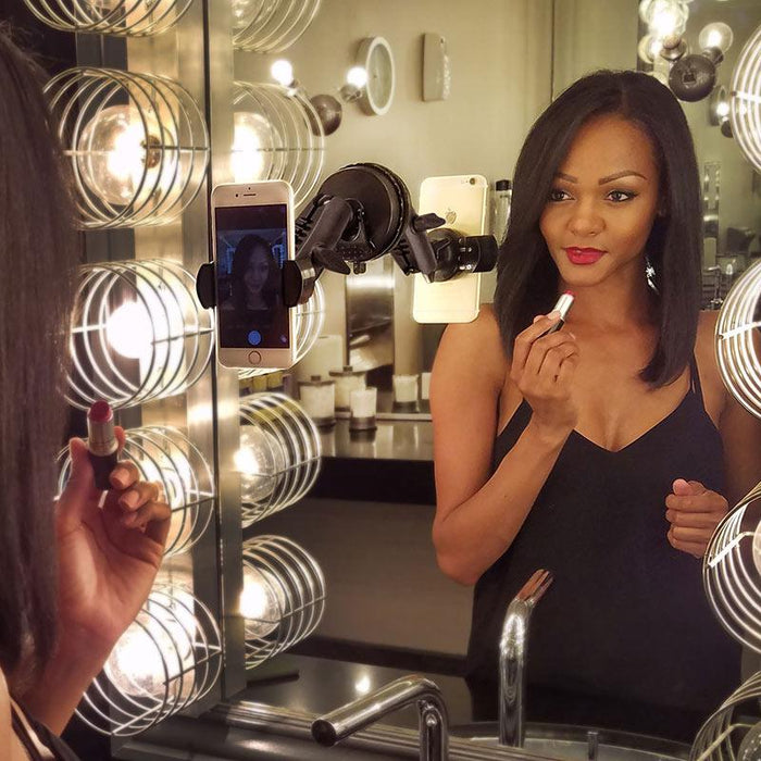 A woman in a black top applies lipstick while looking in a well-lit mirror, her phone filming her reflection using the RoadVise® Phone Holder with Sticky Suction Mount and Shaft.