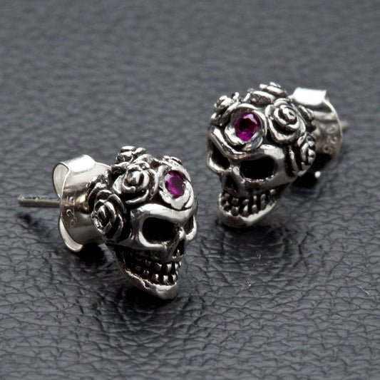 A pair of sterling silver rose skull earrings with brilliant red stone accents shown on a black leather surface.