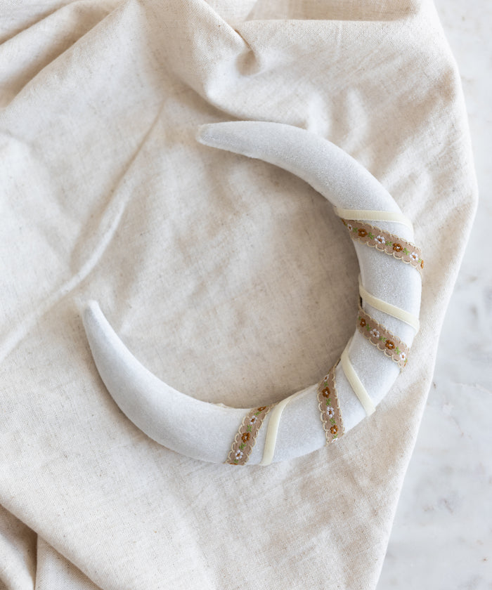 The SAMPLE SALE Headband Swirl White by WALD Berlin, featuring a crescent shape with sequin and ribbon details in white velvet, is displayed on a light beige fabric background.