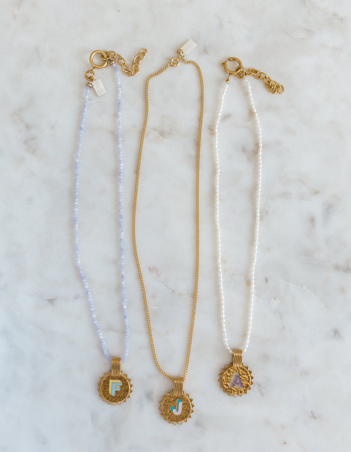 Three WALD Berlin Sample saleSay My Name Gold Necklaces feature gold-plated round initial pendants (F, J, A) with adjustable chains in light blue, gold, and white, elegantly displayed on a marble surface.