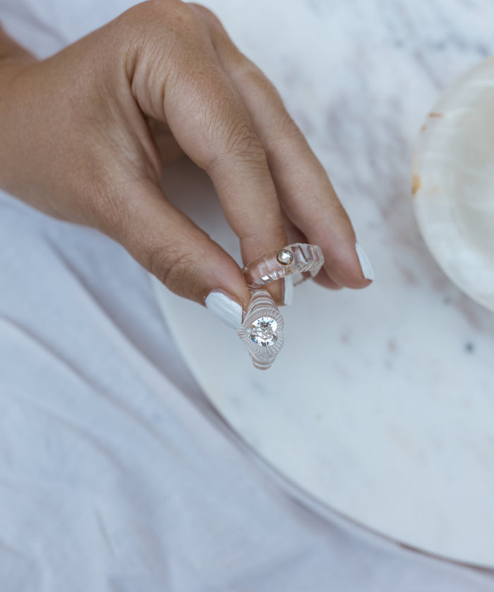 A hand with white polished nails holds WALD Berlin's Shining Star Crystal Ring, showcasing a glamorous pearl adornment and a modern twist with a transparent Swarovski stone, elegantly set over a marble-like surface.