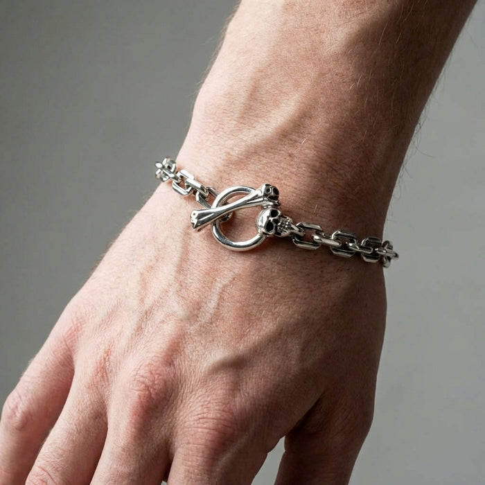 Sterling silver skull T-bar curb link bracelet with toggle clasp featuring skull and crossbones design worn on a male wrist against a neutral gray background