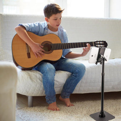 A boy sits barefoot on a white couch, playing an acoustic guitar while looking at a tablet placed on a Slim-Grip® Ultra iPad mini Desk Stand Tablet Holder (17-to-29-Inch Tall), likely following music or instructions.