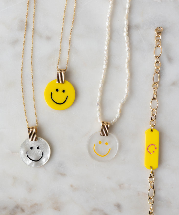 Four pieces of WALD Berlin jewelry on a marble surface: three necklaces, including the Smilie Dude Pearl Medaillon Necklace with smiley face pendants, and one gold chain bracelet with a handmade yellow rectangular charm featuring a smiley face.