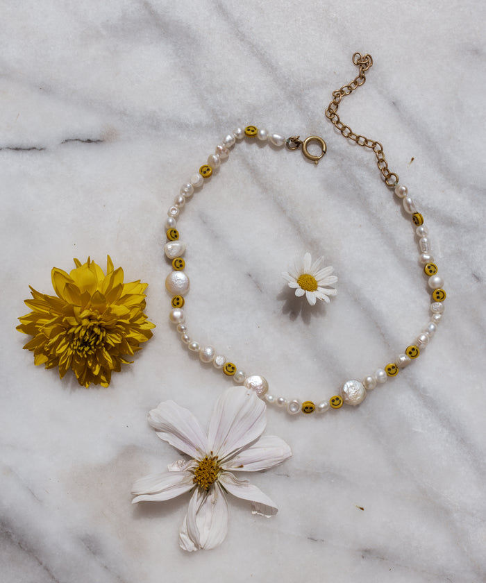 The Smilie Dude Pearl Necklace by WALD Berlin lies elegantly on marble, showcasing sweetwater pearls with smiley beads, surrounded by a vibrant yellow flower and delicate daisy-like blooms.