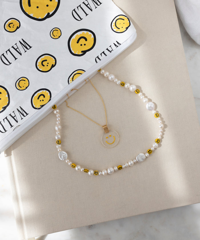 The WALD Berlin Smilie Dude Pearl Necklace, handmade with sweetwater pearls, gold beads, and smiley faces, is displayed beside a gold pendant necklace and a white pouch with yellow smileys and "WILD" text.